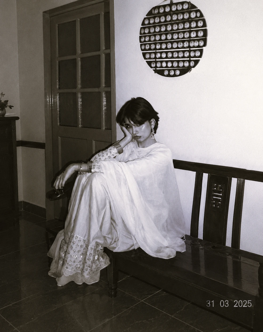 A woman in a white embroidered suit and dupatta sits on a wooden bench in a hallway, her chin resting on her hand.